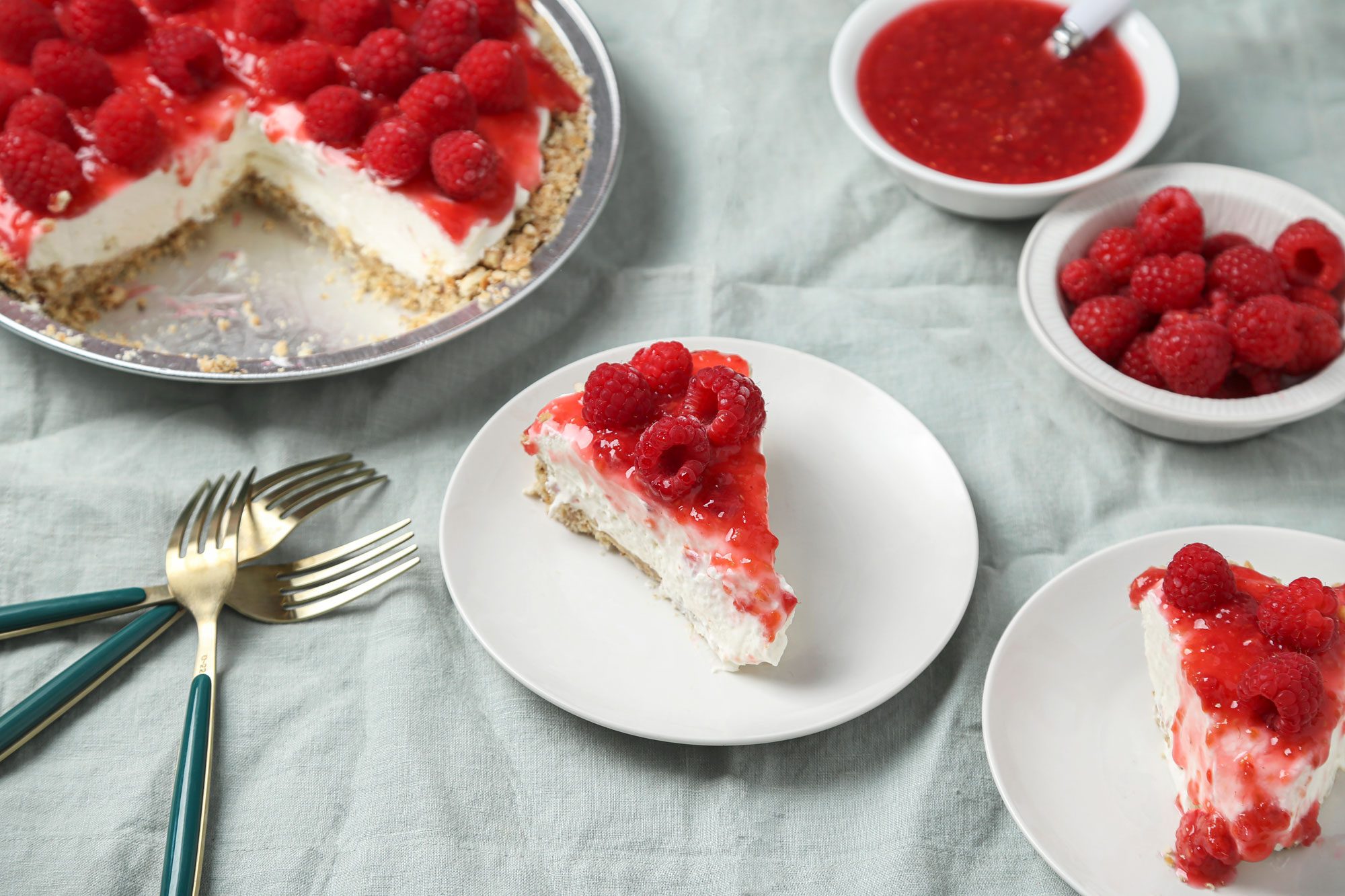 3/4th shot of slice of raspberry topped cheesecake sits on a white plate, with more cheesecake, fresh raspberries, raspberry sauce, and forks nearby on a light blue tablecloth