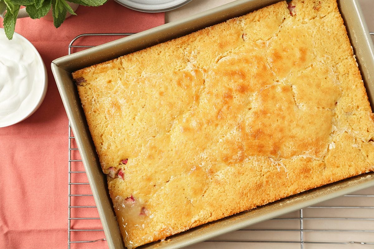 A golden-brown baked cake in a rectangular metal pan rests on a cooling rack, with a bowl of whipped cream and a pink napkin nearby.