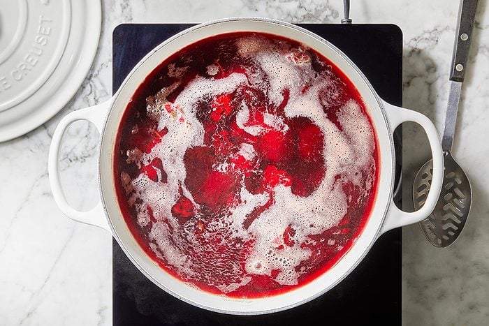 A white pot filled with bubbling red liquid, possibly simmering fruit or berry jam, sits on a stovetop with a slotted spoon nearby.