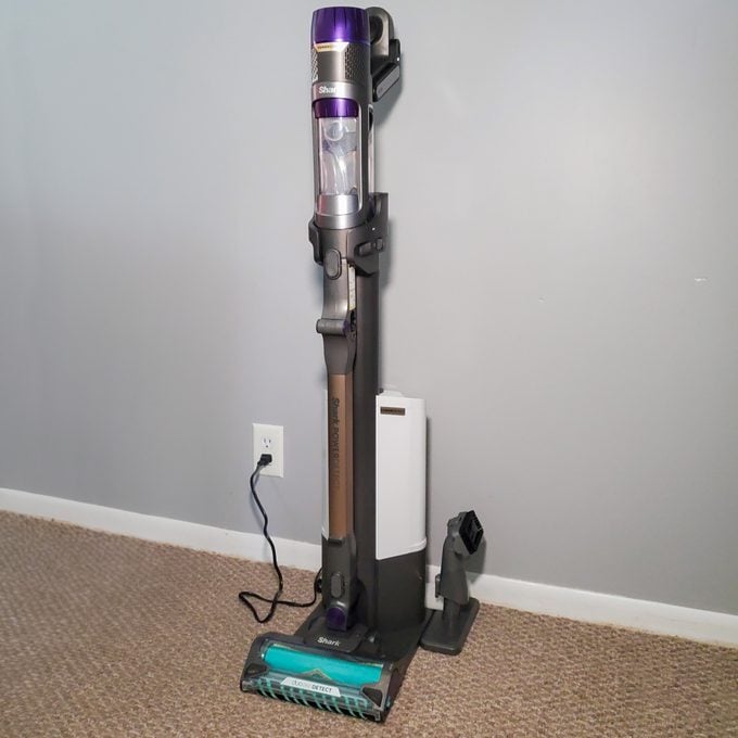 A cordless upright vacuum cleaner with a purple and silver handle is standing on a beige carpet next to a gray wall outlet, with a power cord plugged in.