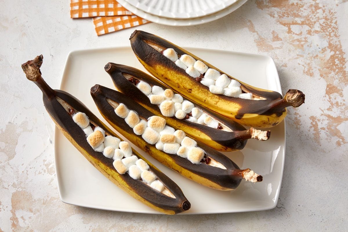 Overhead shot of Banana Boats served on a white plate; empty plates visible on two orange and white checkered napkins; two spoons nearby; all set on a marble surface;