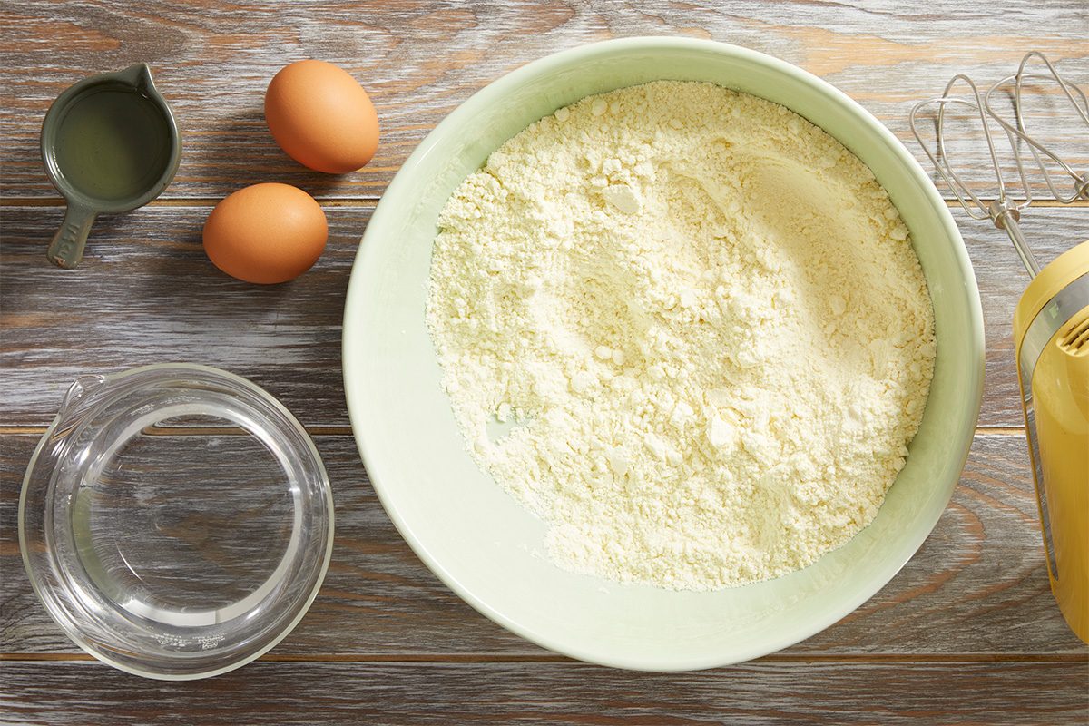 A bowl of flour mixture sits on a wooden surface next to two eggs, a glass measuring cup of water, a small container of oil, and a yellow electric hand mixer.