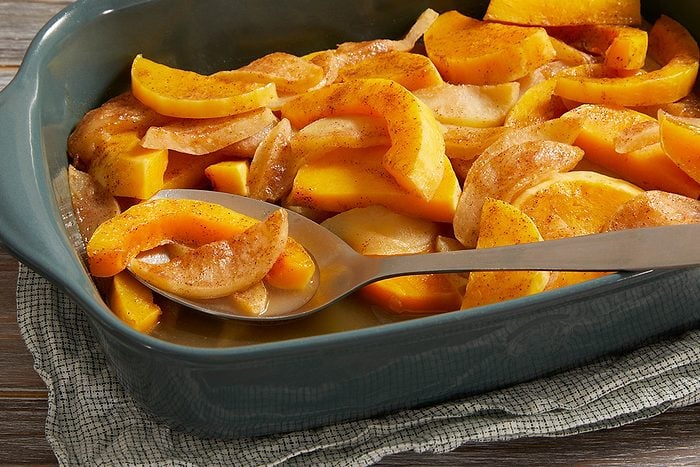 A baking dish filled with sliced yellow squash and onions, seasoned and baked, with a metal serving spoon resting inside. The dish sits on a textured cloth on a wooden surface.