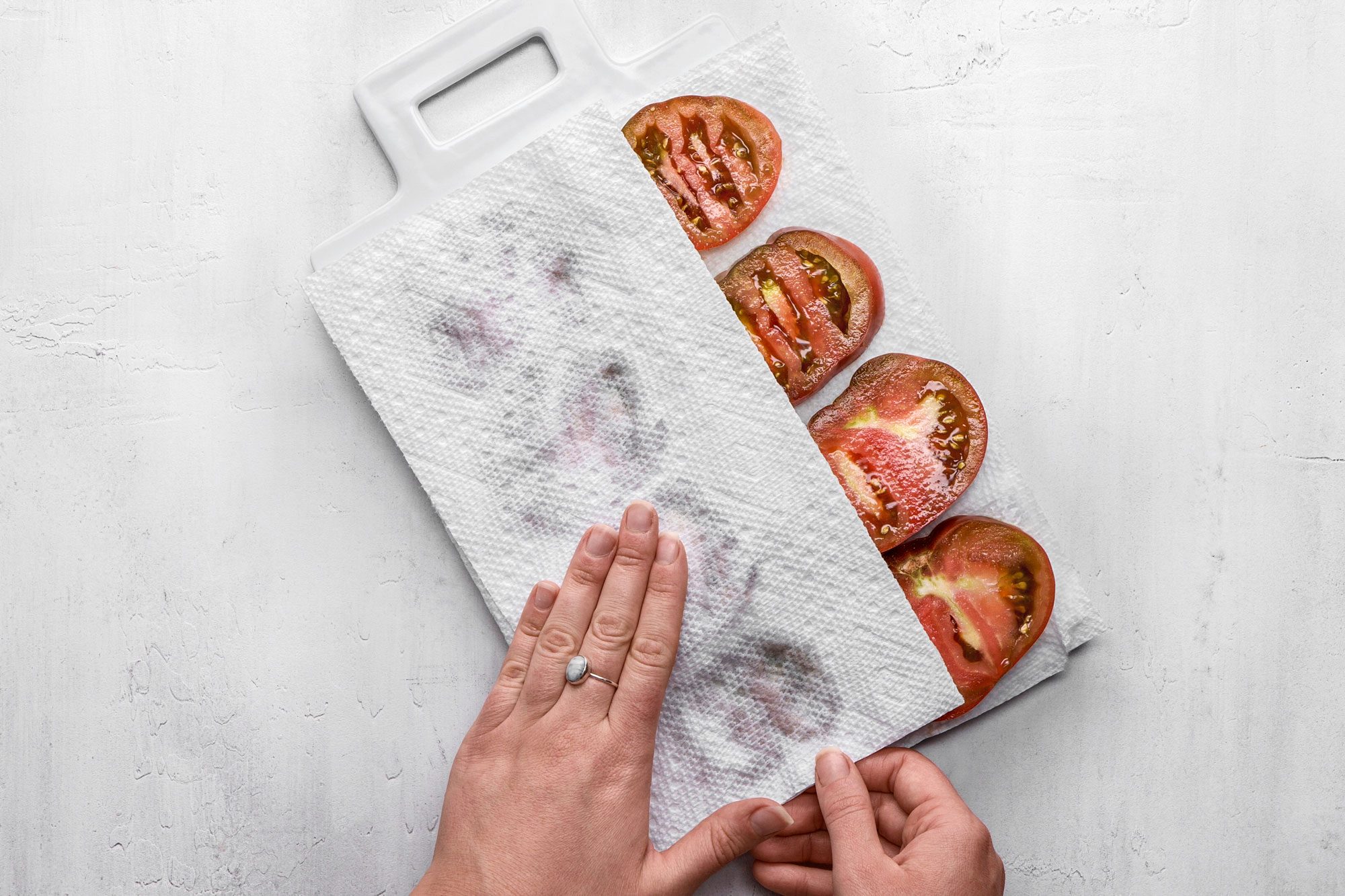 A top shot of a Hands gently pat sliced tomatoes dry using a paper towel placed on a white cutting board; the background is solid white; emphasizing freshness and kitchen preparation