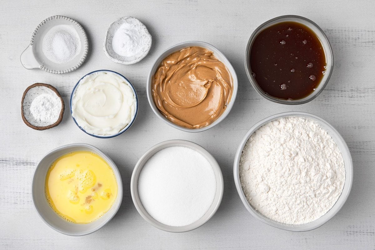 Top Shot of a Eight bowls on a white surface hold flour; sugar; melted butter; yogurt; peanut butter; honey; baking powder; baking soda and salt; organized for baking preparation