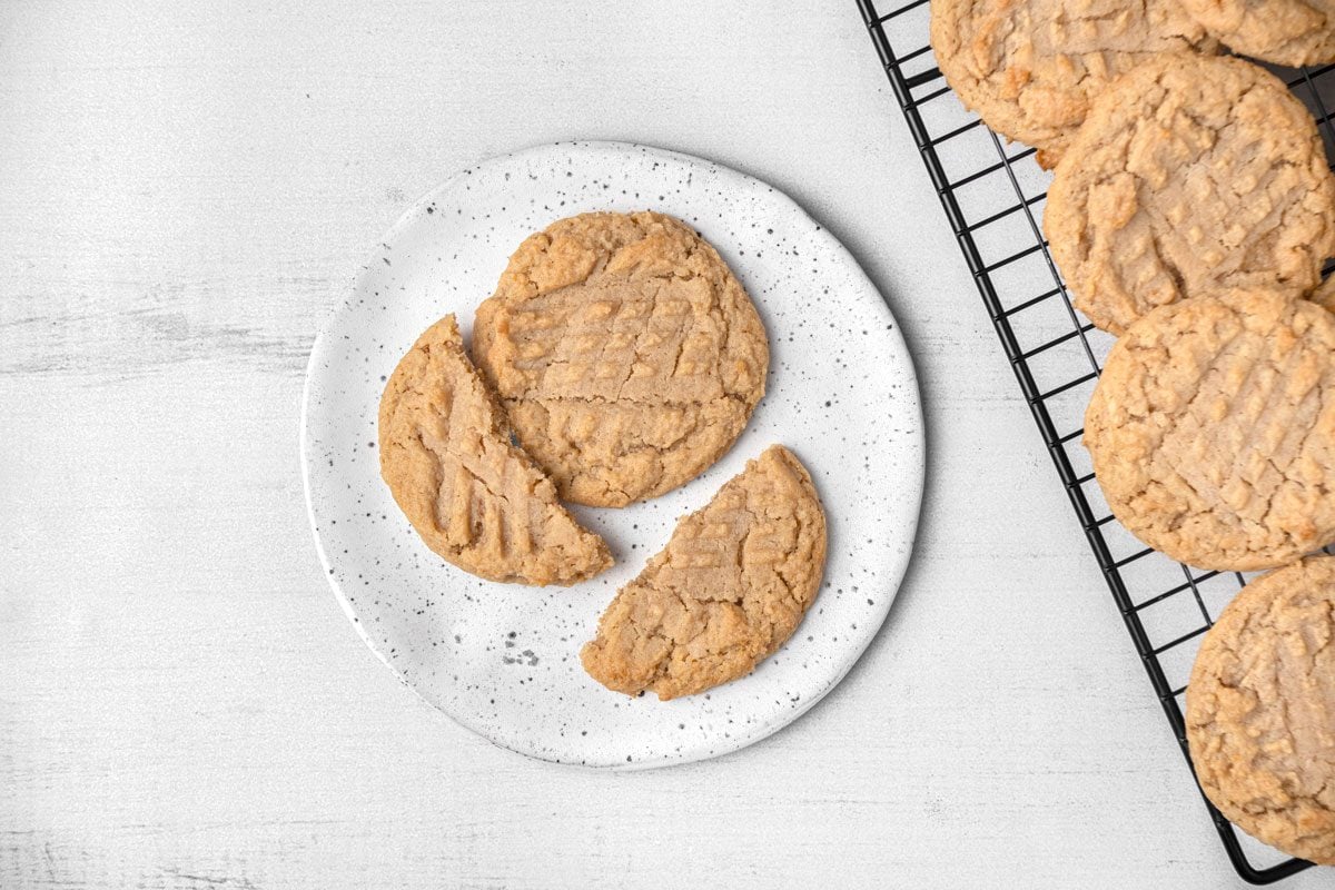Top shot of a white plate holds three whole honey peanut butter cookies and one broken cookie on a white surface; a cooling rack with extra cookies is to the right; the background is minimalistic and bright