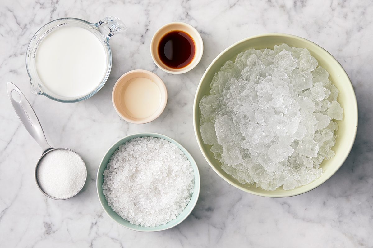 A marble countertop displays bowls and cups with ingredients: a large bowl of crushed ice, a measuring cup of milk, a bowl of shredded coconut, a small cup of sugar, vanilla extract, and condensed milk.