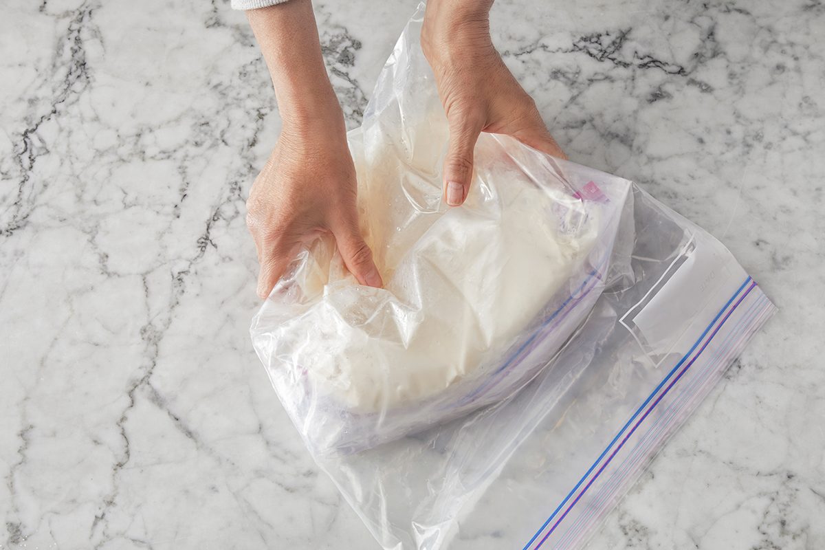 Two hands seal a large plastic zip-top bag filled with a white substance, possibly dough, on a marble countertop.