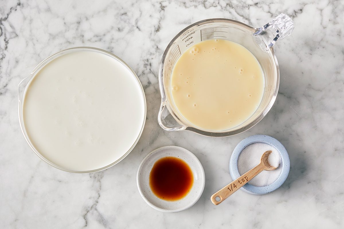 Four ingredients on a marble surface: a bowl of milk, a measuring cup with a creamy liquid, a small dish of vanilla extract, and a small bowl of salt with a wooden spoon.
