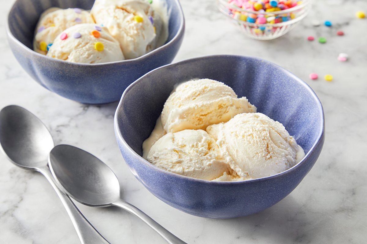 Two blue bowls with vanilla ice cream, one topped with colorful sprinkles, sit on a marble surface. Two metal spoons and a small bowl of sprinkles are nearby.