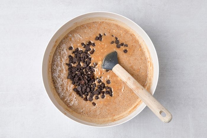 A mixing bowl filled with light brown batter and chocolate chips, with a black spatula resting inside. The bowl is placed on a light-colored surface.