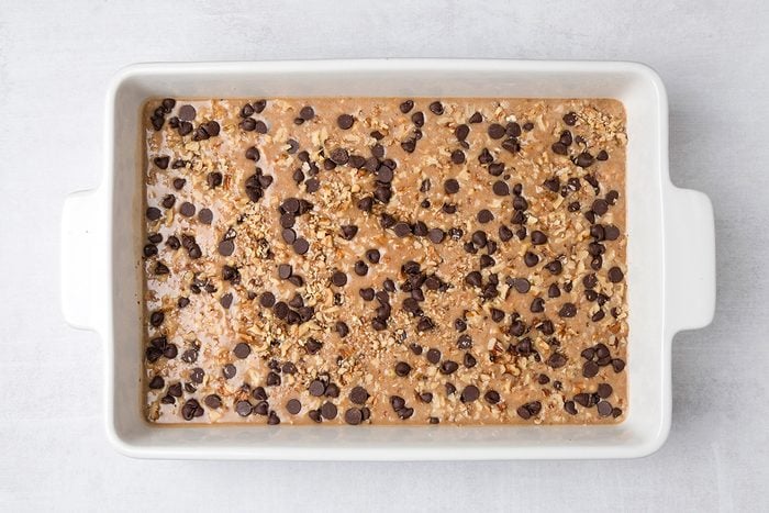 A rectangular white baking dish filled with unbaked batter, topped with chocolate chips and oats, sits on a light-colored surface.