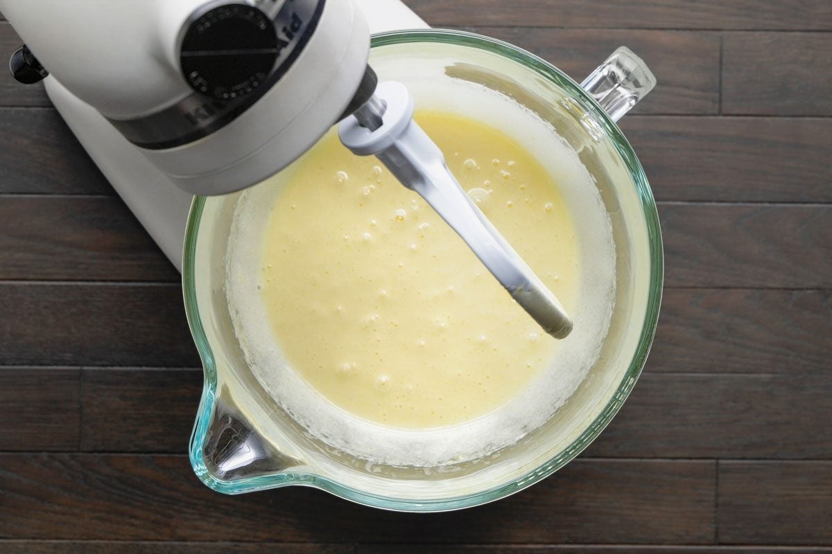 Overhead shot of a large bowl with eggs and sugar being beaten until frothy using a blender; all set on a dark brown wooden surface;