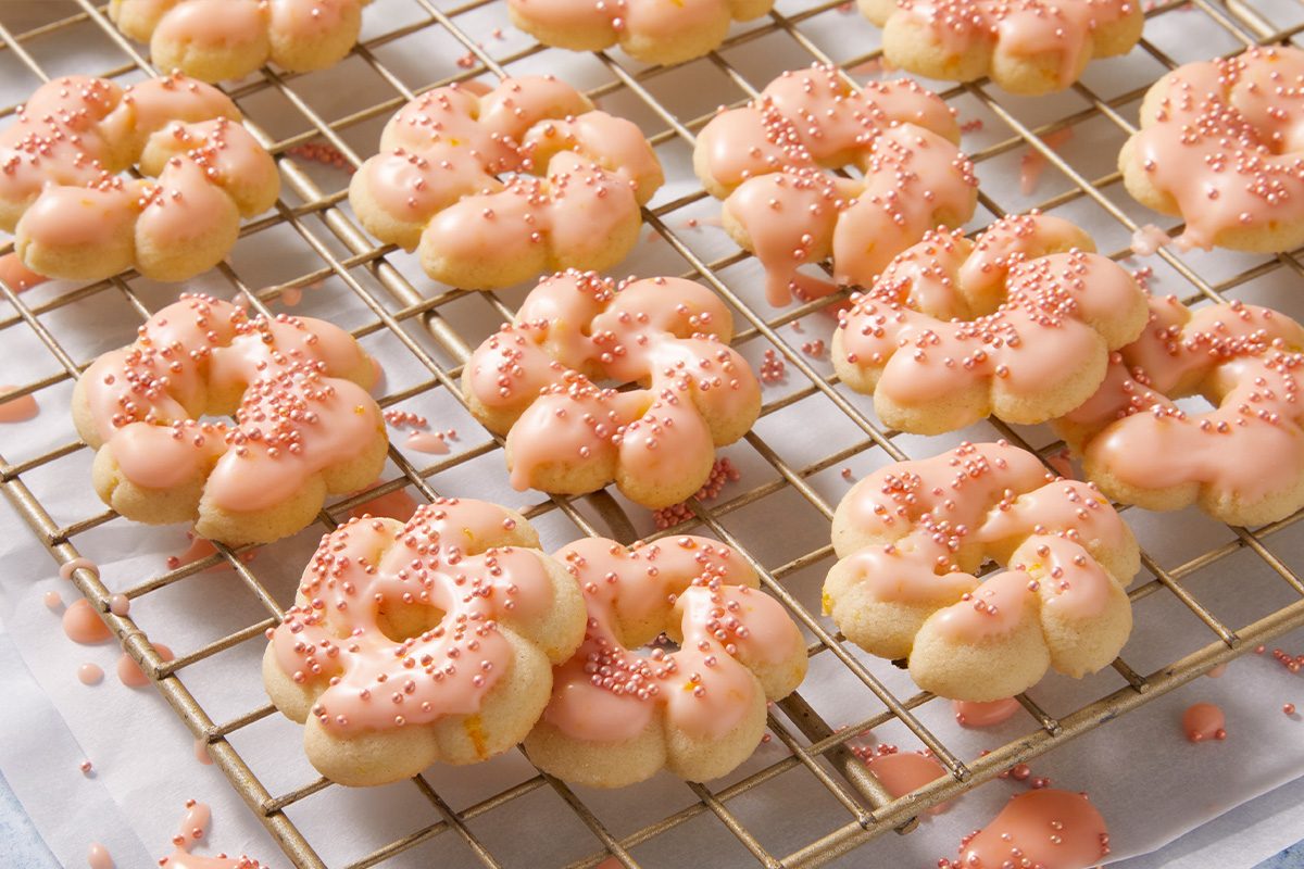 Ring-shaped cookies with pink icing and pink sprinkles sit on a cooling rack. Some icing and sprinkles have scattered onto the surface below. The cookies appear freshly decorated and arranged neatly.