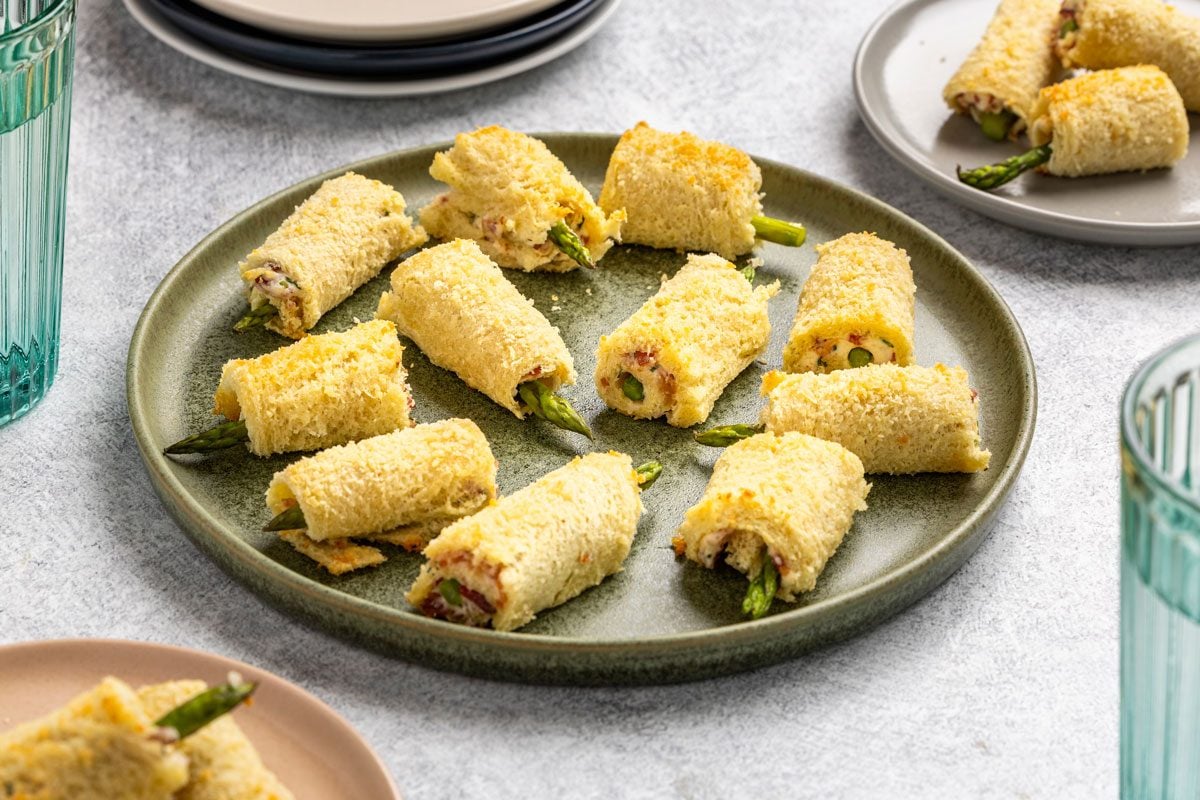 Overhead shot; green plate with rolled bread bites filled with asparagus; filling; surrounded by plates; glasses on a light grey surface