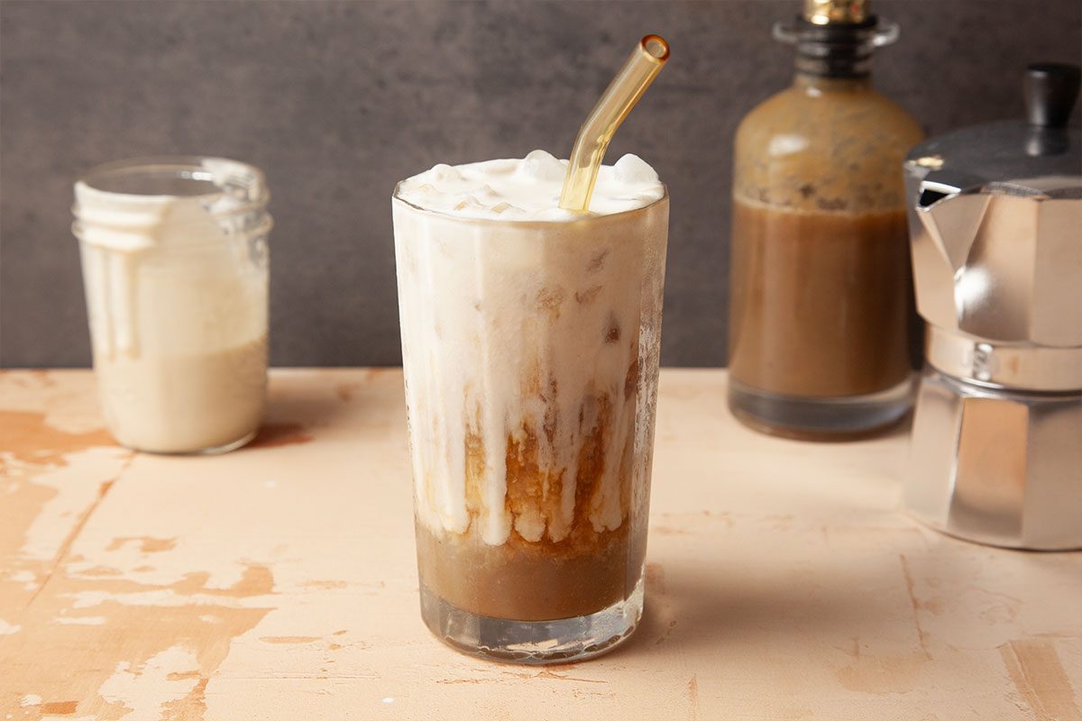 A glass of iced coffee topped with creamy milk and a glass straw, sitting on a light wooden surface. In the background, there are jars and a moka pot slightly out of focus.
