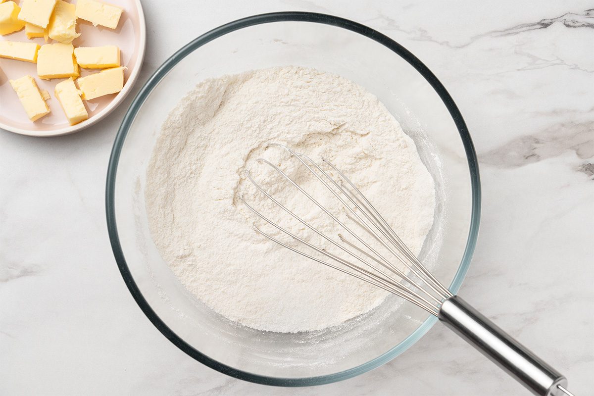 A glass bowl filled with flour and a metal whisk on a marble surface. A plate with cubed butter sits nearby in the upper left corner.