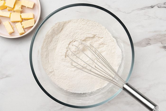 A glass bowl filled with flour and a metal whisk on a marble surface. A plate with cubed butter sits nearby in the upper left corner.