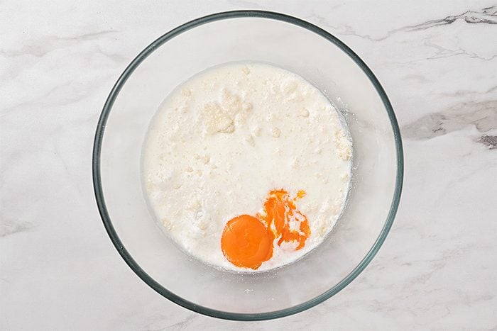 A glass bowl on a marble surface contains milk, flour, and two cracked eggs with visible yolks, ready to be mixed together.
