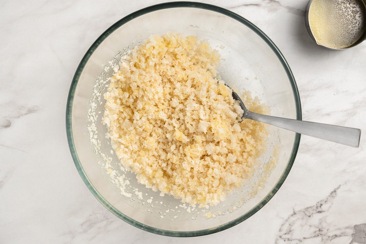 A glass bowl filled with a mixture of creamed butter and sugar sits on a marble surface, with a metal spoon resting inside the bowl. Another small bowl with butter residue is nearby.