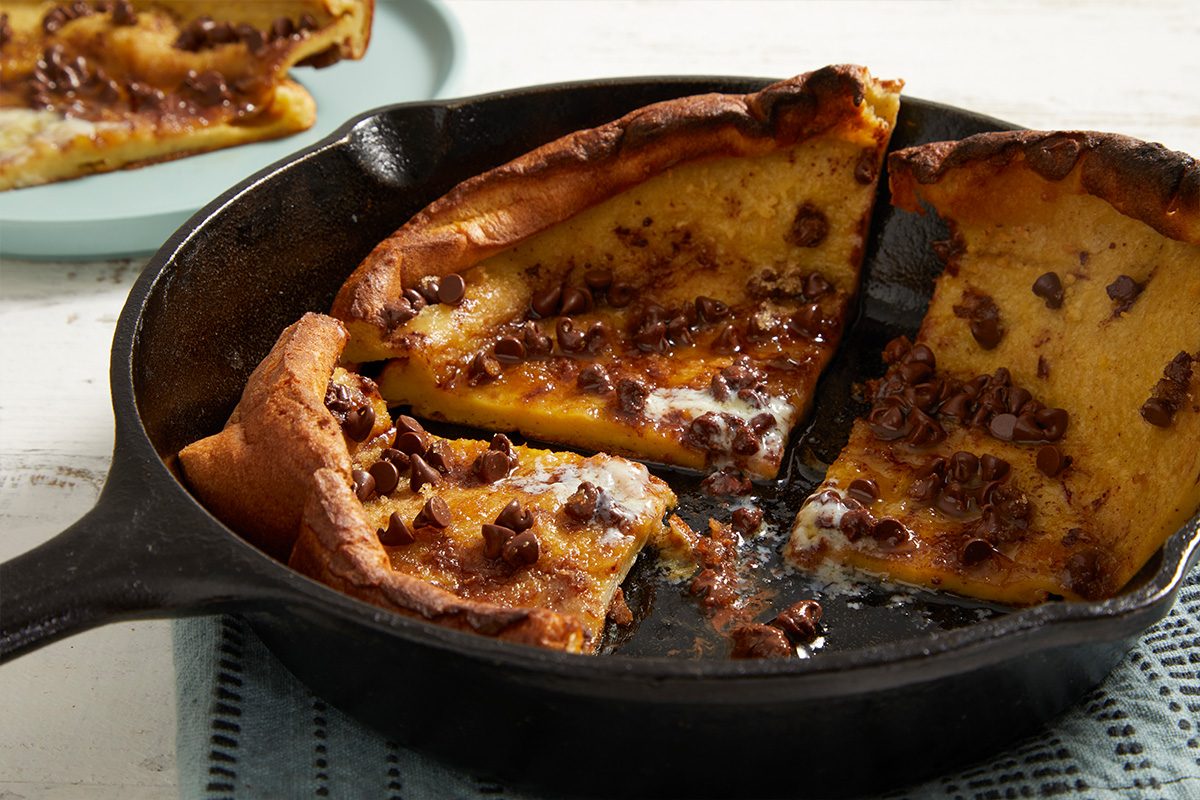 A chocolate chip Dutch baby pancake is sliced in a cast iron skillet, with a portion served on a plate in the background. The pancake is golden brown with melted chocolate throughout.