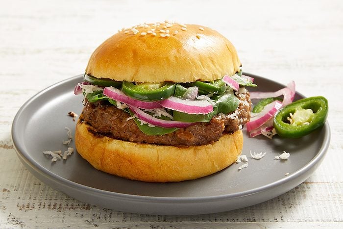 A hamburger on a gray plate, topped with sliced red onions, jalapeños, lettuce, and cheese in a sesame seed bun, with extra jalapeño slices on the side.