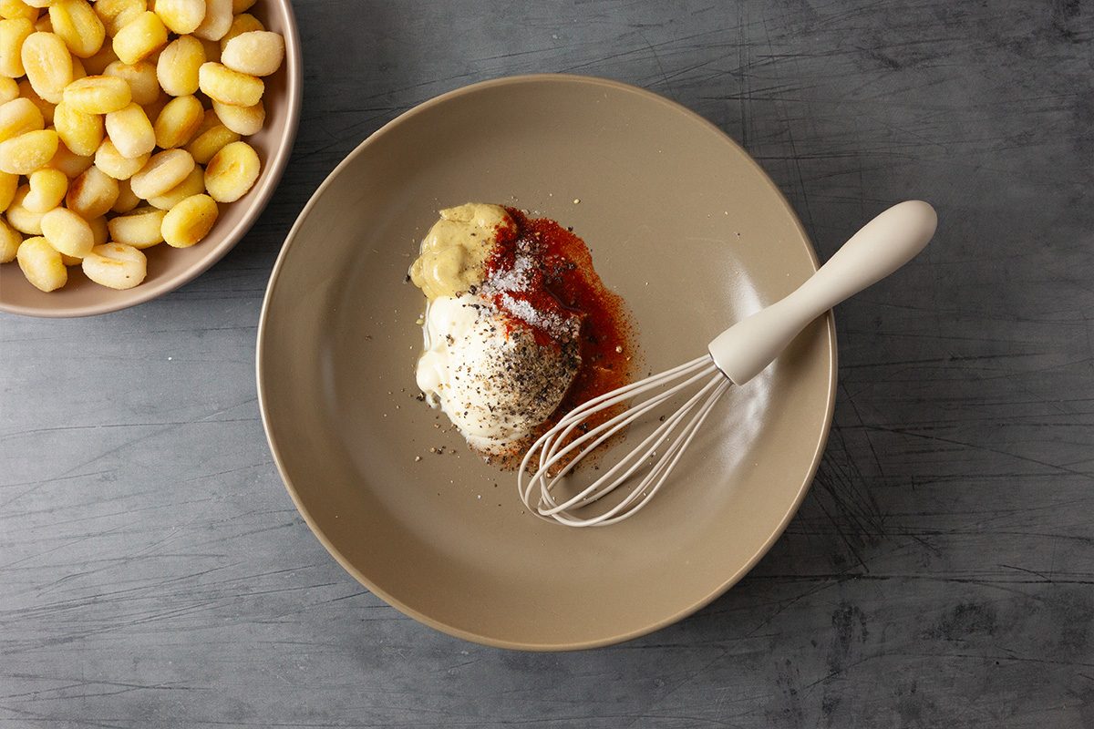 A beige plate with mayonnaise, ketchup, grated cheese, and spices sits next to a whisk. A bowl of uncooked gnocchi is nearby on a gray tabletop.