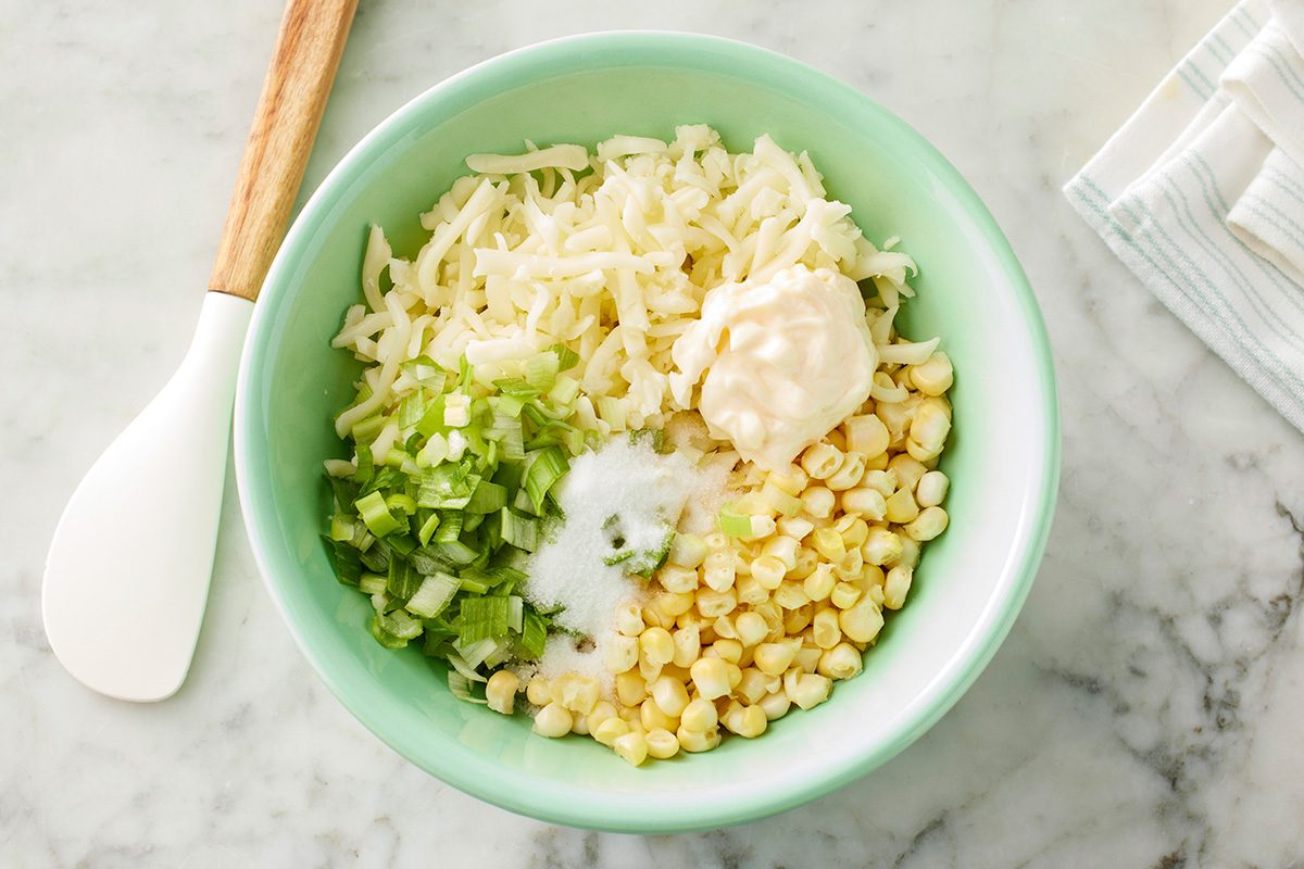 This is a top-down shot showing a green bowl on a marble surface filled with shredded cheese; corn kernels; chopped green onions; mayonnaise; and salt, A white spatula and striped cloth are placed beside the bowl for preparation