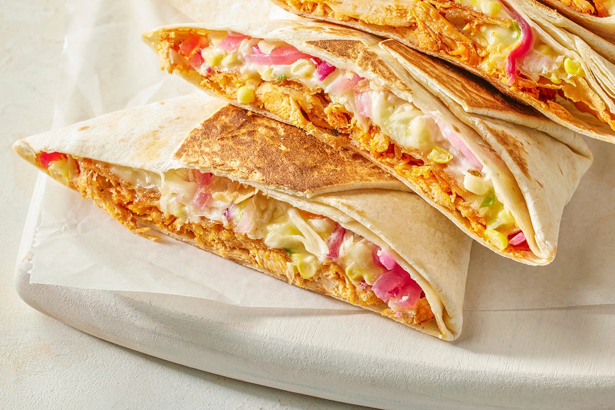 This is a close-up shot. Three tortilla wraps, halved and stacked; filled with shredded chicken; lettuce; and pickled onions; rest on parchment paper atop a light-colored wooden board, creating an inviting food presentation