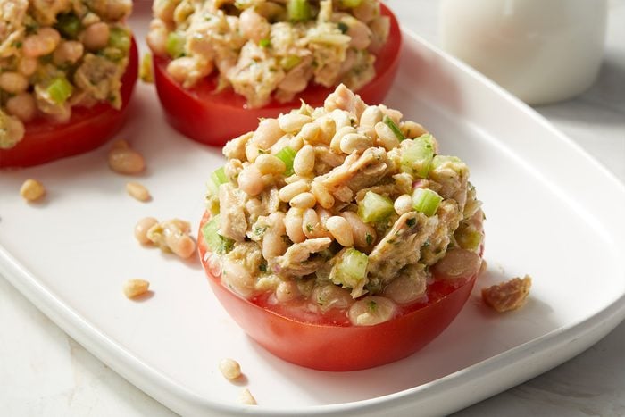 A halved tomato topped with a mixture of tuna, white beans, celery, and seasonings is served on a white plate, creating a fresh and colorful appetizer.