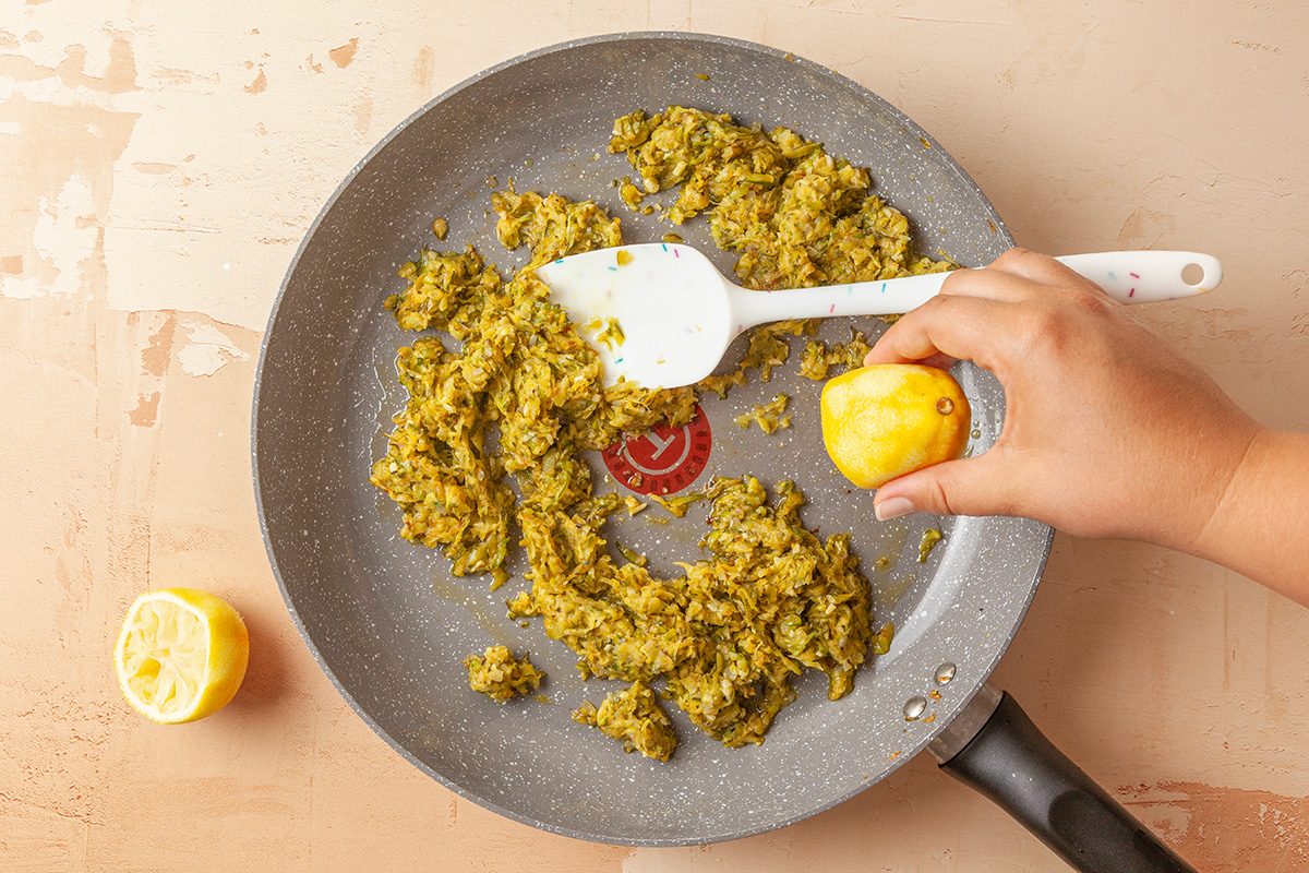 This is a close-up shot; a hand squeezes a lemon over a frying pan with greenish-yellow vegetables; a white spatula rests inside; another lemon half is on the beige countertop