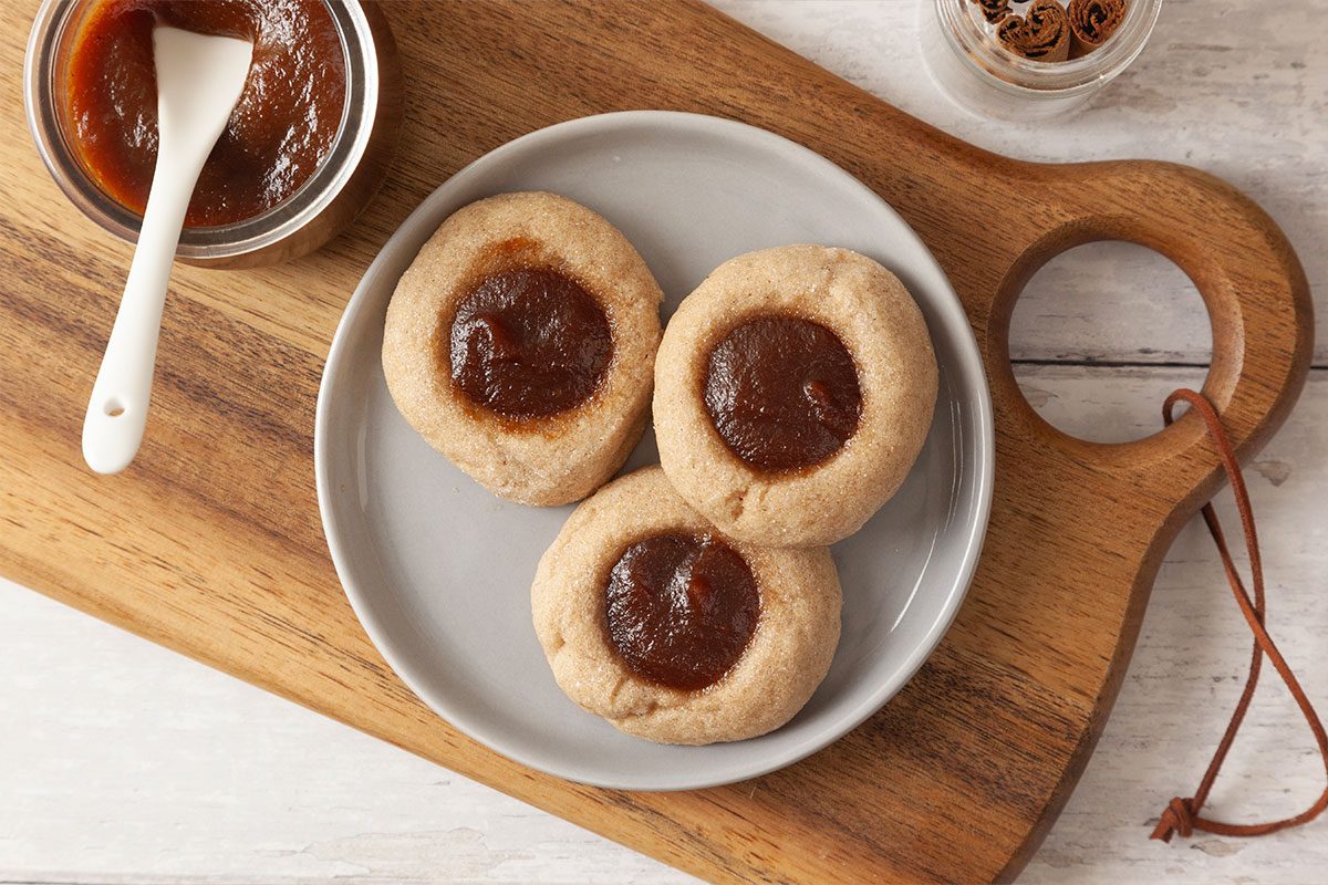 Three thumbprint cookies filled with brown jam are arranged on a round plate, which sits on a wooden serving board. A small bowl of jam with a white spoon is nearby.