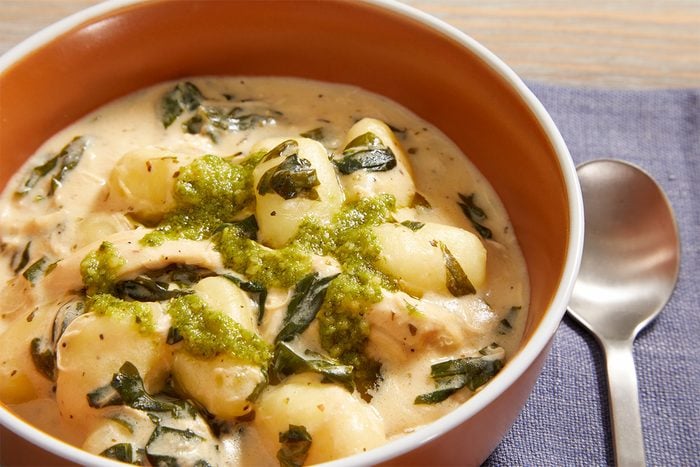 A bowl of creamy gnocchi soup with spinach and shredded chicken, topped with a swirl of green pesto, sits on a purple napkin beside a silver spoon.