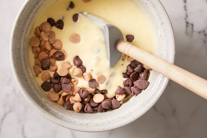 A mixing bowl with creamy batter, chocolate chips, and peanut butter chips being stirred with a gray spatula on a marble surface.