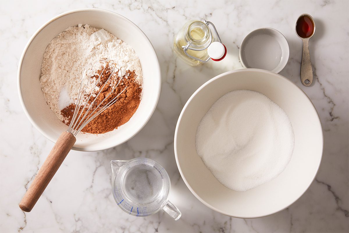 Bowls on a marble surface containing flour and cocoa, sugar, a whisk, a glass of water, a small jar of oil, a dish of vanilla extract, and a bowl of water, arranged for baking preparation.