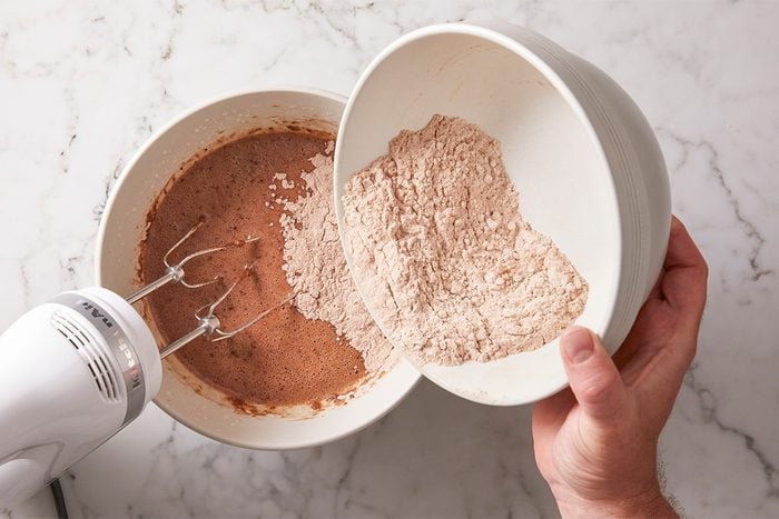 A person pours a bowl of cocoa powder mixture into another bowl with chocolate batter, using an electric hand mixer on a marble countertop.