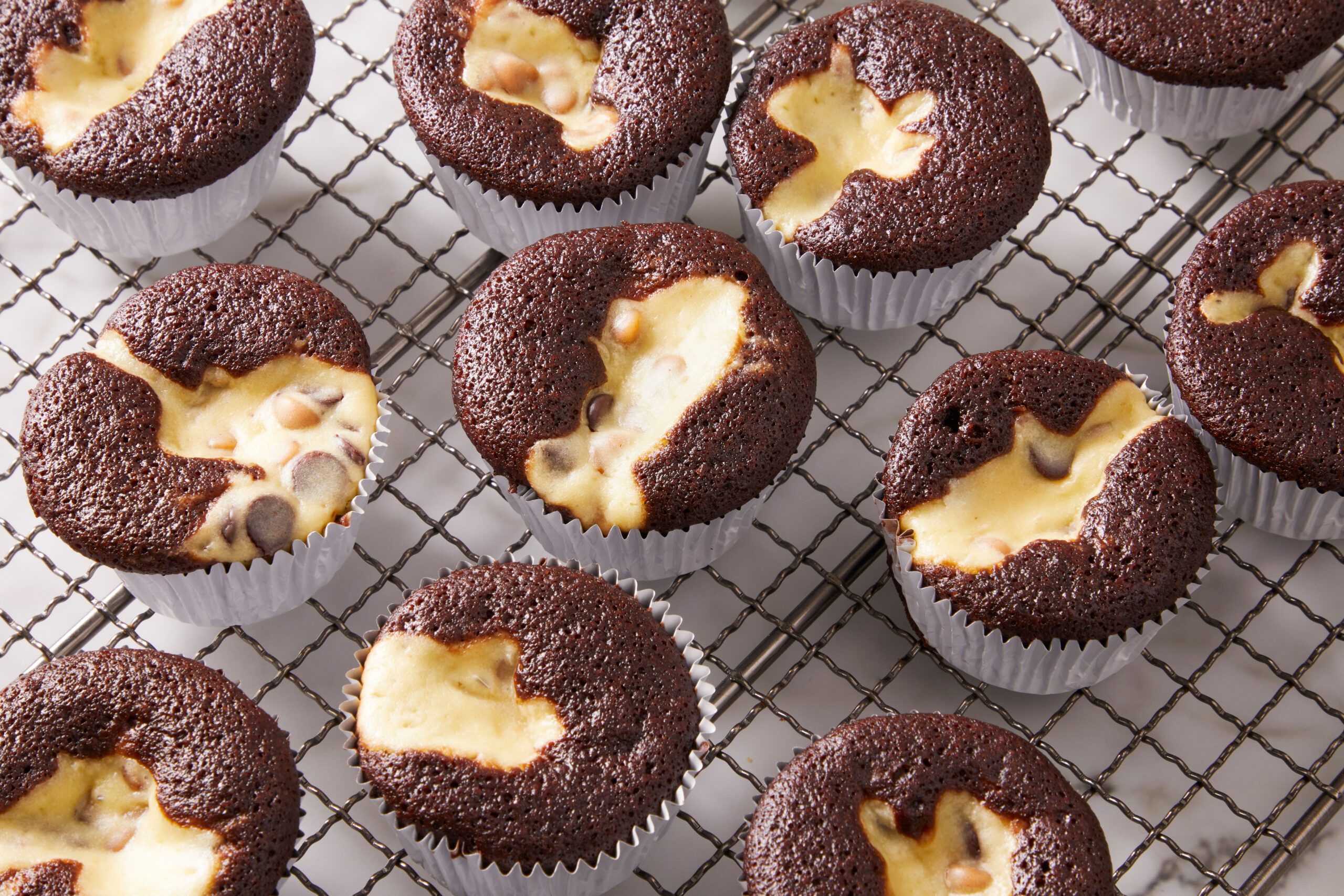 Chocolate cupcakes with creamy, cheesecake-like centers and chocolate chips are cooling on a wire rack, each in a white paper liner.