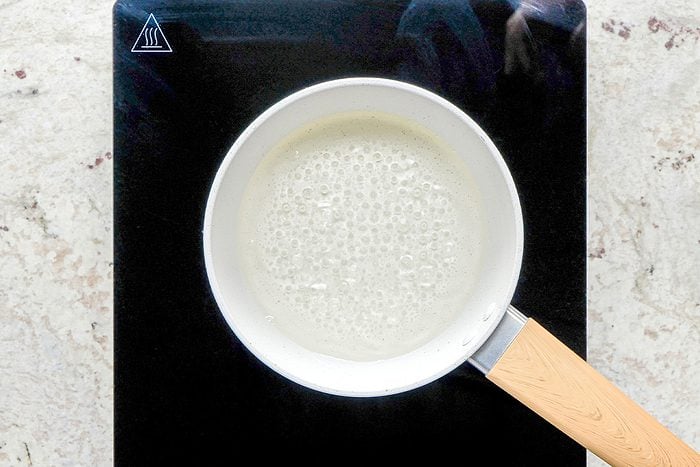 A white saucepan with a light wooden handle is heating a bubbling liquid on a black stovetop, viewed from above. The stovetop is set on a light-colored countertop.
