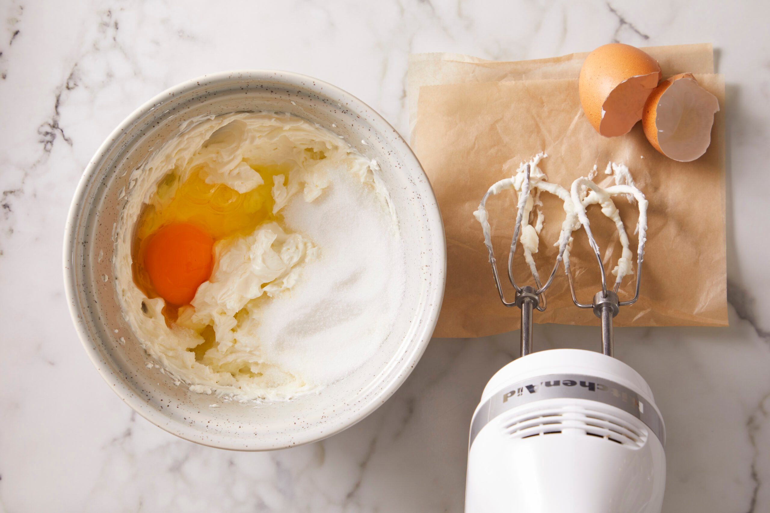 A mixing bowl with creamed butter, sugar, and an egg, next to a hand mixer with beaters and cracked eggshells on parchment paper, all on a marble countertop.