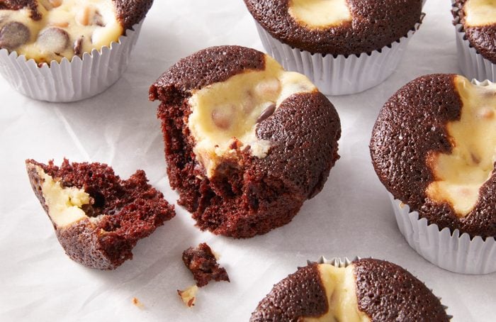 Chocolate cupcakes with creamy cheesecake filling and chocolate chips, some in white paper liners, one broken open to show the moist texture inside, all on a white surface.