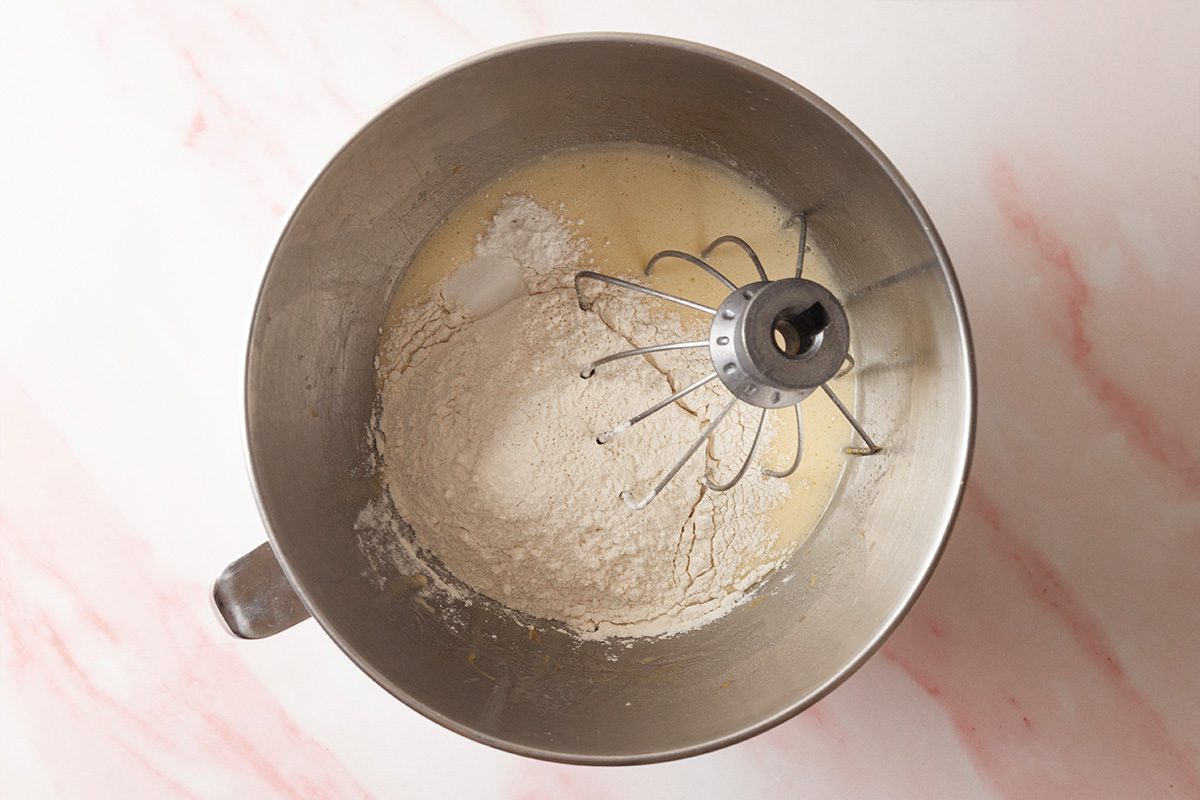 A metal mixing bowl with a whisk attachment contains flour and liquid ingredients, ready to be mixed, on a light pink and white marbled countertop.