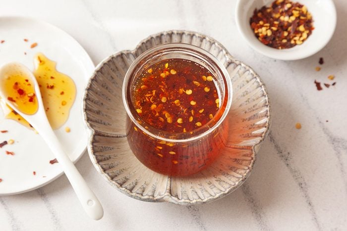 A glass jar filled with chili oil sits on a decorative plate, surrounded by a white spoon with chili flakes and oil, a white plate with oil stains, and a small bowl of more chili flakes on a marble surface.