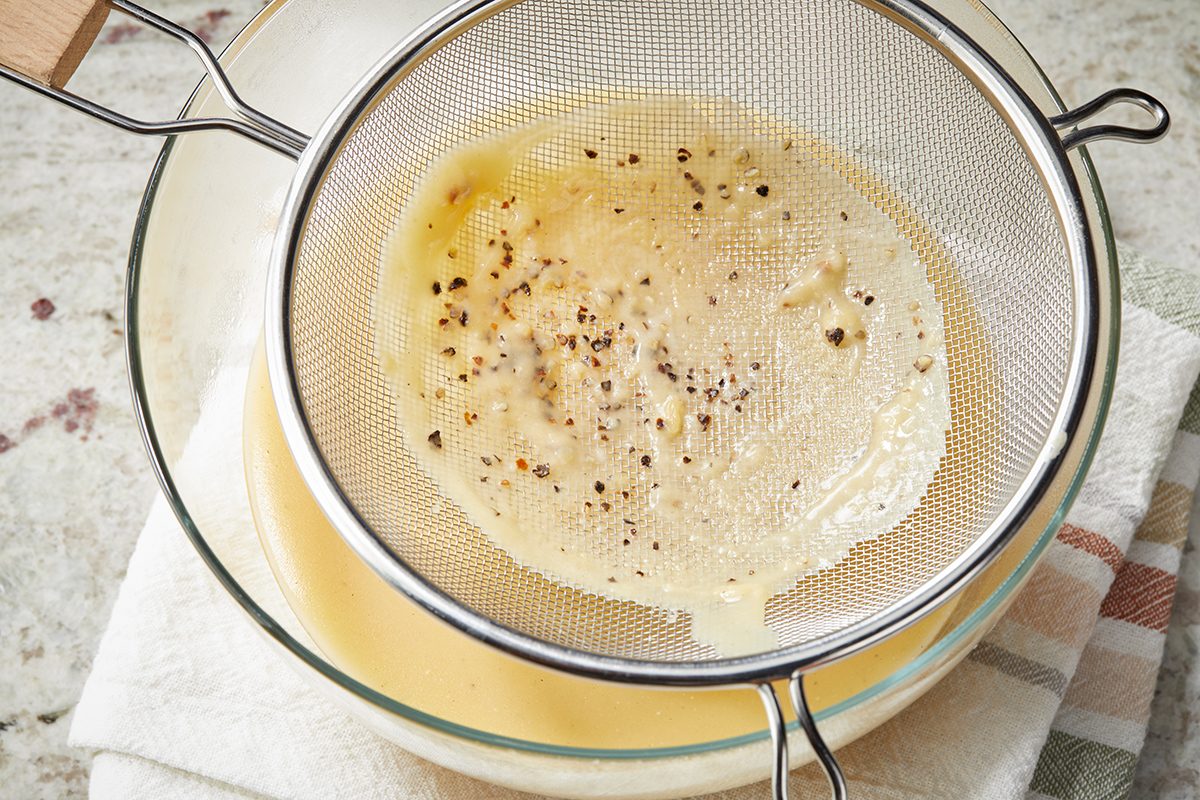 A fine mesh strainer sits over a glass bowl, filtering a creamy liquid with ground black pepper and solids caught in the strainer. The bowl rests on a white towel with thin colored stripes.