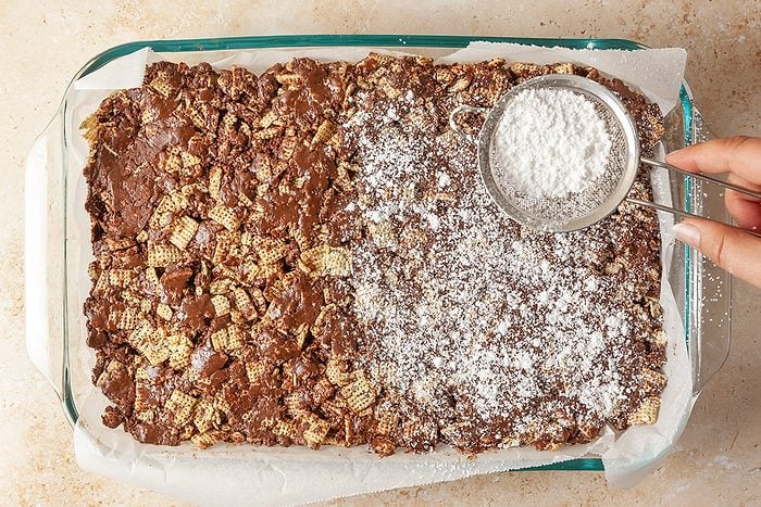 Overhead shot of a hand sprinkling powdered sugar over a chocolate cereal bar mixture in a glass baking dish; with half of the dessert covered in sugar while the other half remains plain