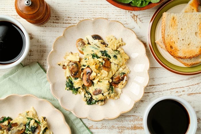 A plate of scrambled eggs with mushrooms and spinach on a cream-colored plate, next to slices of toasted bread, two cups of coffee, and a pepper grinder on a rustic white table.
