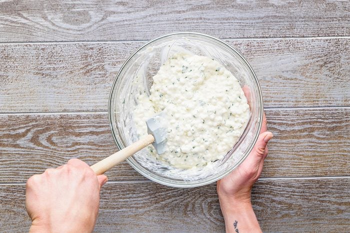 A person holding a glass bowl filled with a creamy, chunky mixture and stirring it with a white spatula, on a wooden surface.