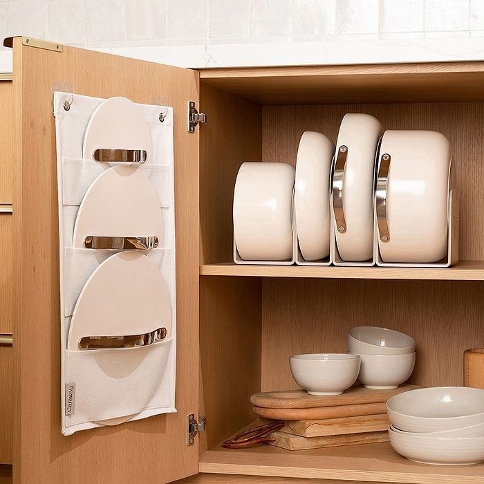 An organized kitchen cabinet with pots and lids neatly stored on shelves; pan lids are also held in a hanging rack attached to the inside of the cabinet door. Bowls and cutting boards are arranged on the bottom shelf.