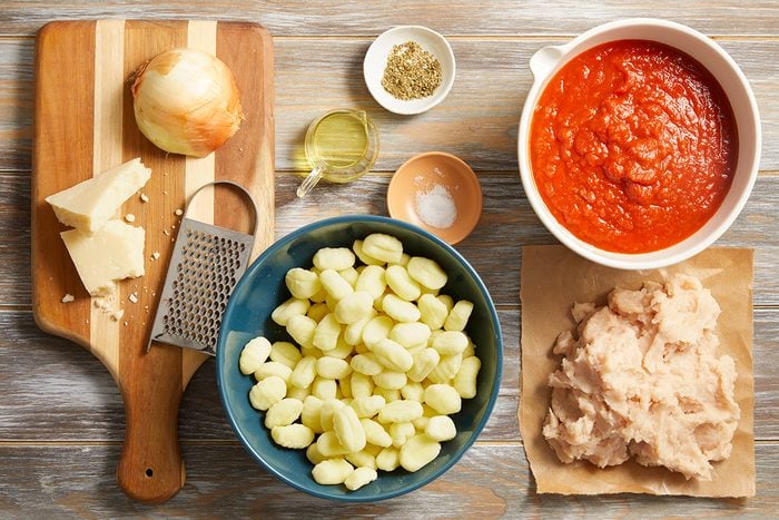 Ingredients for a meal arranged on a table: gnocchi in a bowl, ground meat on paper, tomato sauce in a bowl, an onion, parmesan cheese, a grater, olive oil, salt, and dried herbs.