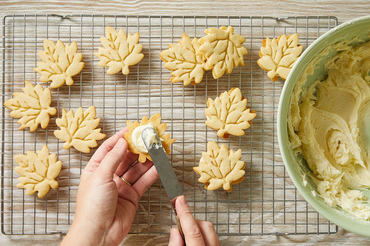 Overhead shot of a person spreads frosting on a leaf-shaped cookie with a spatula over a cooling rack holding other cookies and a large green bowl of frosting sits nearby on a wooden surface
