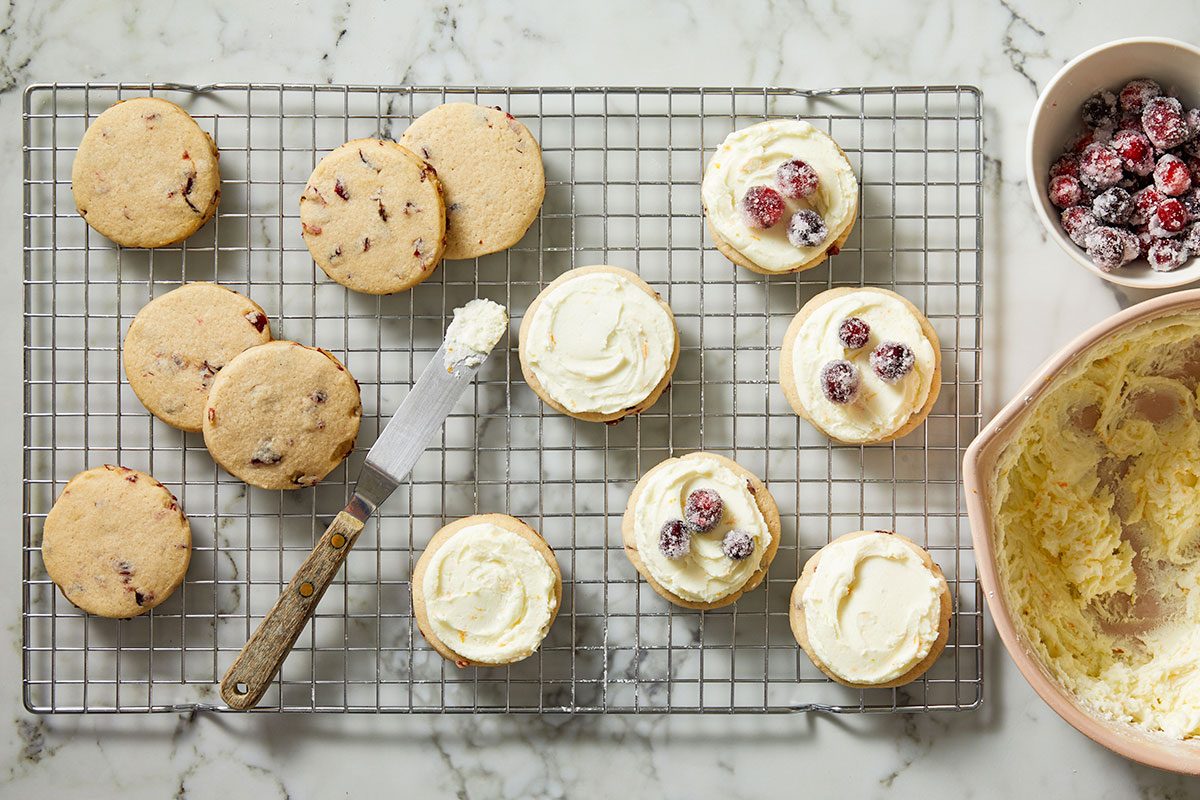 Overhead shot of baked cookies cooling on a wire rack beside a bowl of frosting and sugared cranberries.